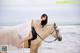 A woman in a white dress riding a white horse on the beach.